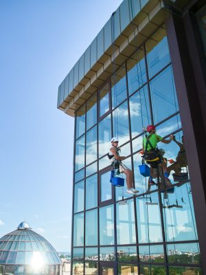 Industrial mountaineering. Male washers hanging on ropes and using cleaning tools while washing building windows. Two men window cleaners in safety helmets working together at high-rise building.